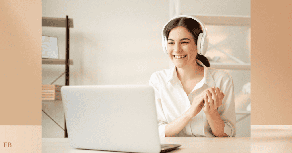 A smiling woman wearing headphones is sitting at a desk, engaged in a video call on her laptop. She appears to be clapping or gesturing positively, with a neatly organized shelf in the background.