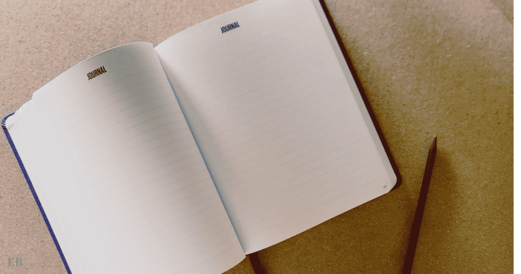 An open journal with the word "JOURNAL" printed at the top of each blank, lined page, accompanied by a sharp pencil resting on a textured brown surface, suggesting a setting prepared for writing or note-taking.
